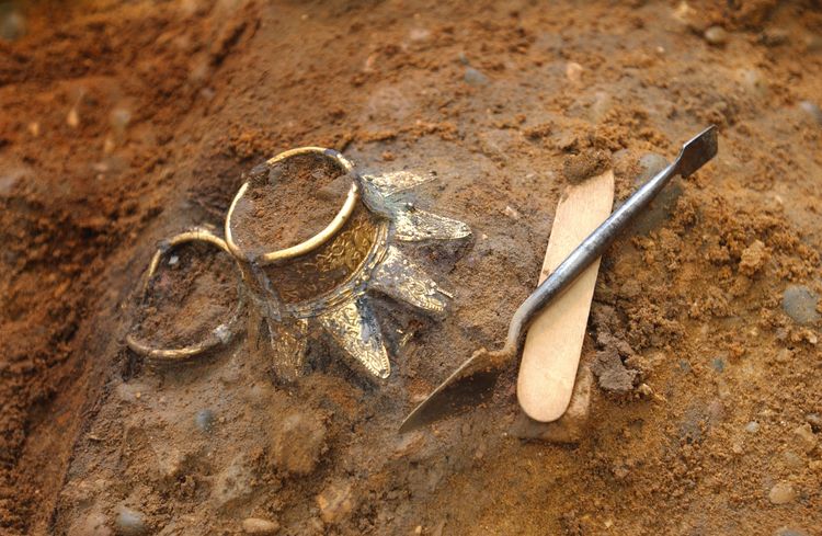 Prittlewell image of two finds, cleaned, but in situ.  They are circular  at the top before continuing into a cylindrical shape with a flared base.  They appear to be made out of gold and there is a trowel next to them for scale.