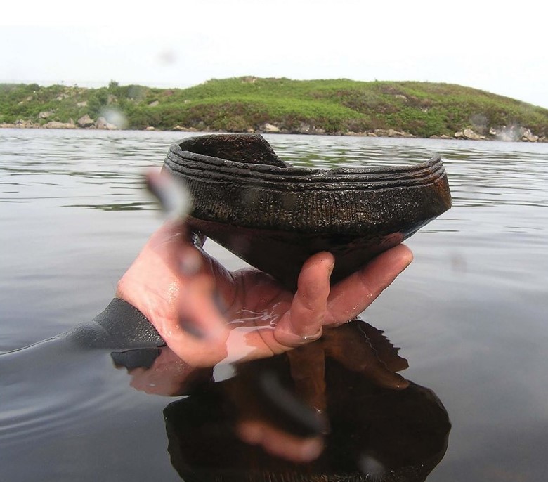 Figure 6. Unstan vessel recovered from loch bed near a crannog in Lewis, Outer Hebrides (photograph by C. Murray, 2012).  A diver's hand holds the diamond shaped vessel just above the water's surface.