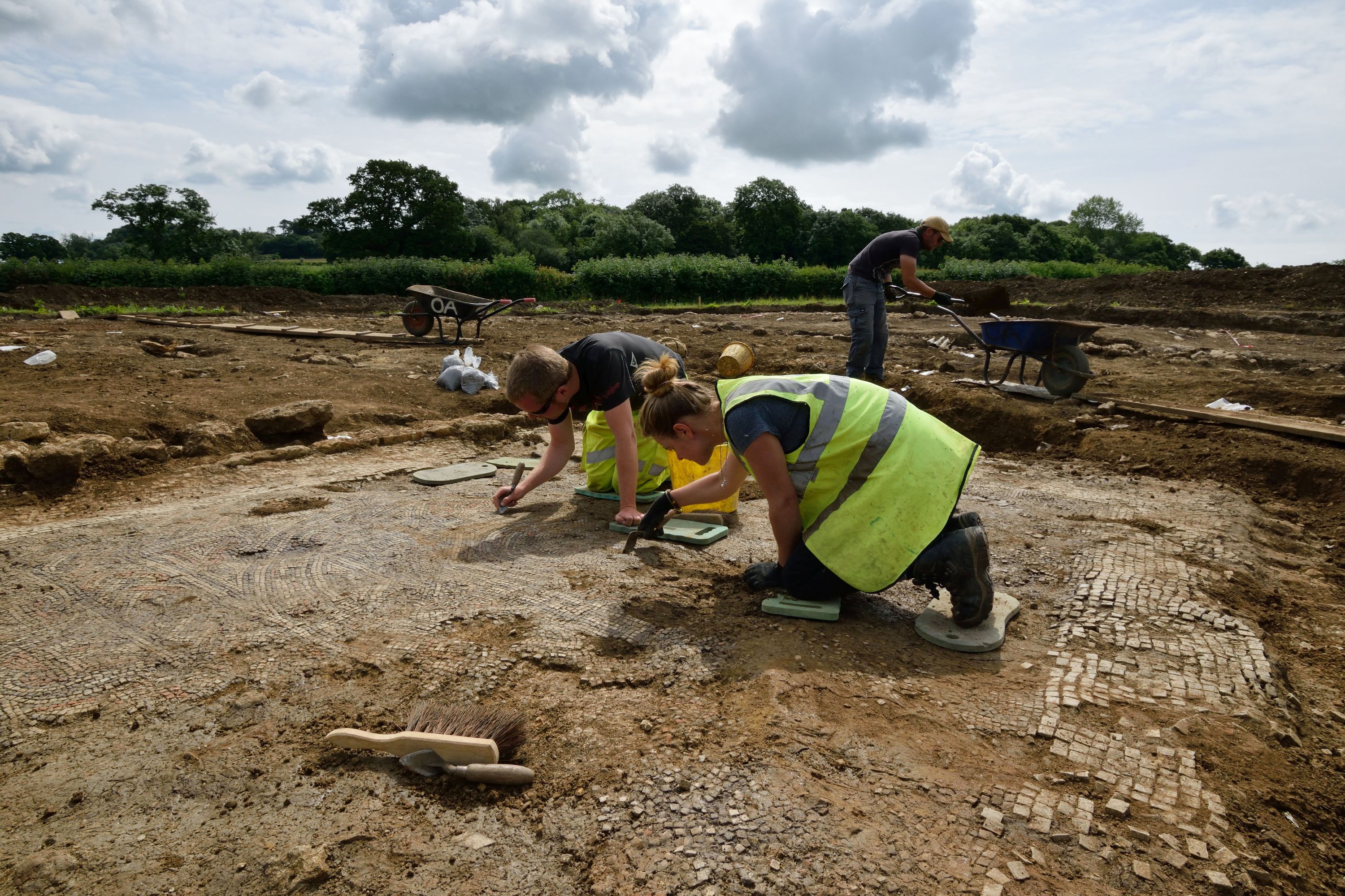 Photograph of two archaeologists on kneelers use their trowels to reveal a mosaic at a Roman villa.