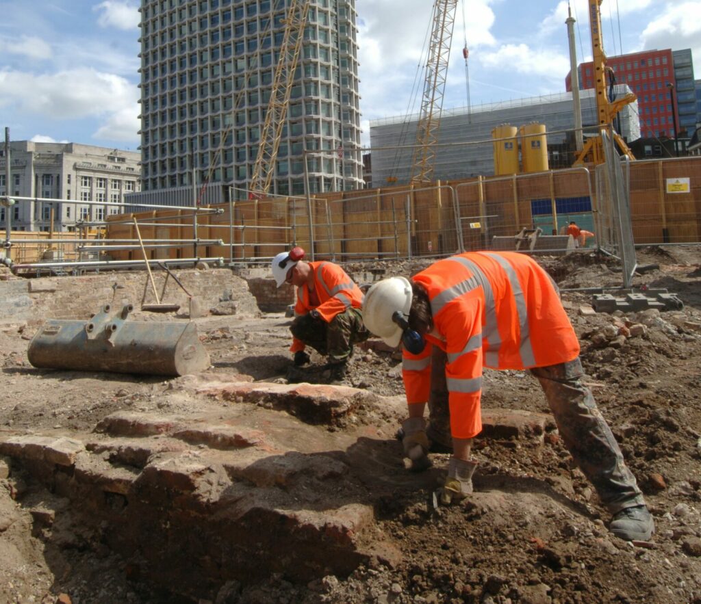 Two people undertaking an archaeological excavation in the shadow of a tall building in London
