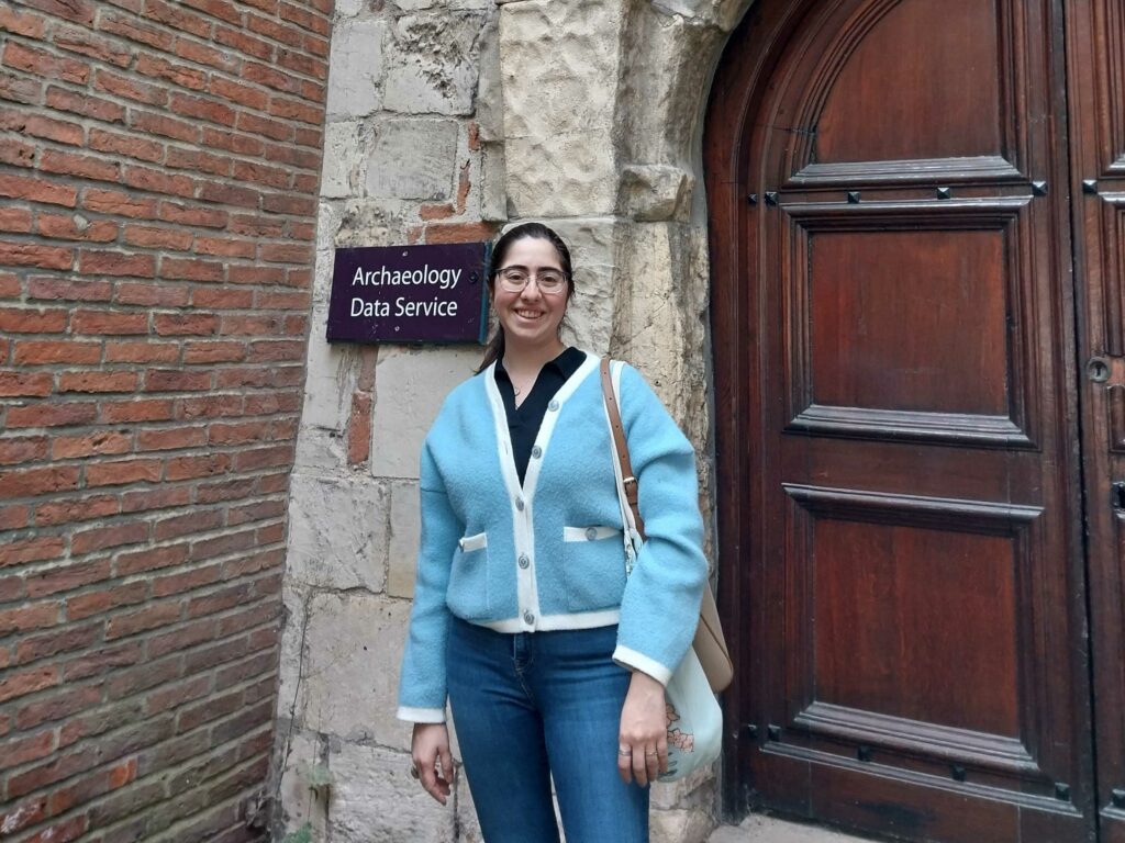 A photo of Burcu wearing a blue cardigan outside the Archaeology Data Service Office