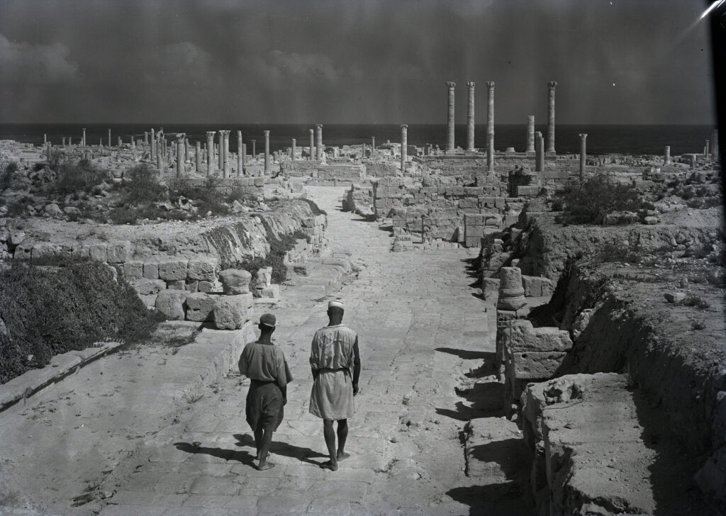 Black and white photo showing two men walking with their backs to the camera. The forum can be seen in the background