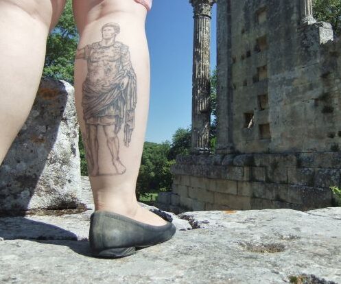 An image of a person with a tattoo of a roman on their leg, with roman ruins in the background