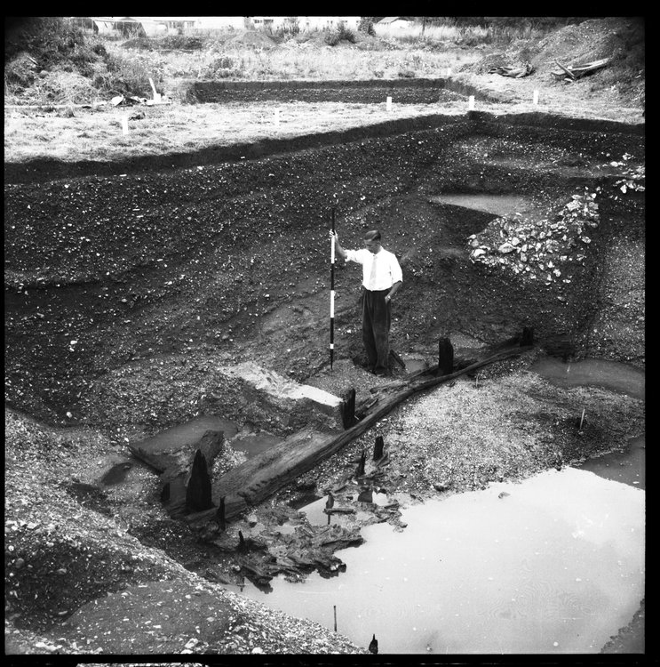 Black and white image showing a excavation with a scale stick and archaeologist for scale