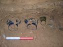 Thumbnail of Glass squat jars <33>, <34>, <35> & <36> (<36> crushed and in fragments at right hand side), all found on their sides adjoinging the east wall of the chamber.