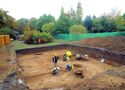 Thumbnail of Excavation work in the shrubbery (background) with the burial chamber just beginning to appear in the centre and an iron age ditch visible in the left foreground